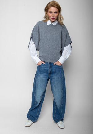 Blonde vrouw staat met handen in de zakken, draagt een wit overhemd onder een grijs kortärmelig trui, wijde blauwe spijkerbroek en witte sneakers.
