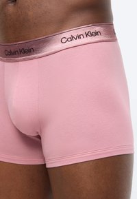 Mies pukeutuneena vaaleanpunaisiin Calvin Kleinin bokserialushousuihin, joissa on metallinhohtoinen vyötärönauha, kuvattuna vyötäröstä reiden puoliväliin yksiväristä taustaa vasten.