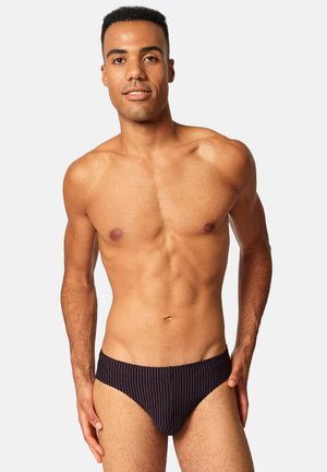 Muskelöser Mann mit kurzen Haaren trägt schwarze, fein gestreifte Slips, steht vor hellem, einfarbigem Hintergrund und lächelt sanft.