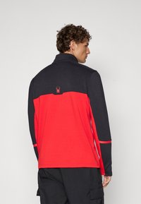 Heren langslavige pullover in zwart en rood, met een hoge kraag, contrasterende kleurvlakken en een klein spinnenlogo op de achterkant.