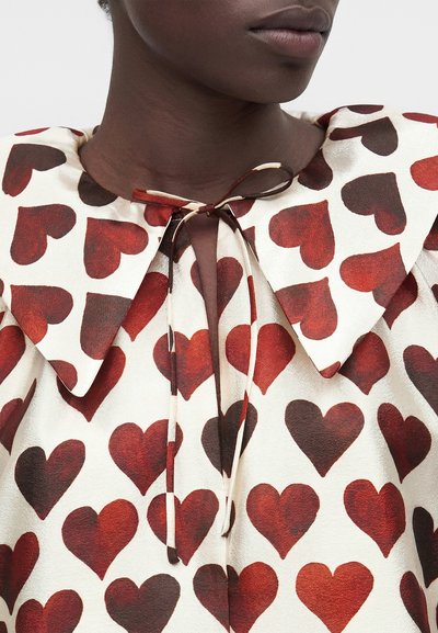 Κοντινό πλάνο ενός ατόμου με σκουρόχρωμο δέρμα που φοράει λευκό μπλουζάκι με κόκκινα σχέδια καρδιών και δεμένη φιόγκο στον γιακά.