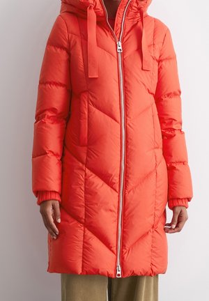 Long manteau matelassé orange avec capuche, deux poches avant et fermeture éclair centrale argentée, porté avec un pantalon en velours côtelé couleur beige.