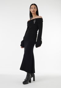 Pull noir en maille, décolleté épaules nues, avec une texture côtelée et des manches larges, associé à une jupe longue noire ajustée et des bottes à plateformes. 