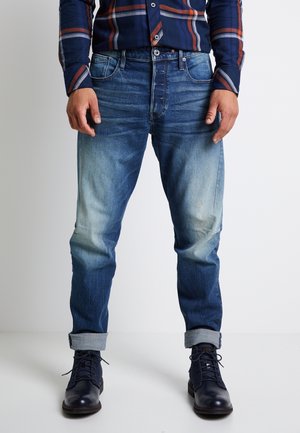 Mann trägt ausgewaschene blaue Jeans mit hochgekrempelten Bündchen, dunkelblaue Schnürstiefel und ein marineblau kariertes Hemd, steht vor weißem Hintergrund.
