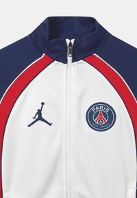 Chaqueta de pista blanca con acentos en navy y rojo, que cuenta con una cremallera, un logo de Jumpman y un emblema circular del Paris Saint-Germain en el pecho.