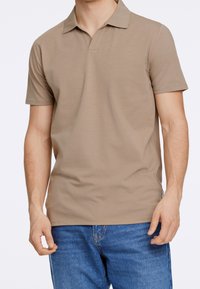 Muž v béžovém polo tričku s krátkým rukávem, které má na sobě, a modrých džínách, stojí na bílém pozadí.