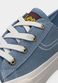 Ανοιχτό μπλε τζιν αθλητικό παπούτσι με χοντρά λευκά κορδόνια, μεταλλικά ρουλεμάν με το λογότυπο "Lee 1889" και εμφανές κίτρινο και κόκκινο υφασμάτινο λογότυπο στη γλώσσα.