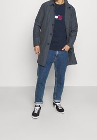 Grå knäppjacka över en marinblå sweatshirt med logotyp, kombinerad med upprullade blå jeans och svarta sneakers. Slät textur, avslappnad passform.