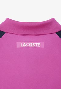 Rosa polo med ribbad krage och marinblå axelaccenter. Har en Lacoste-logotyp på baksidan och textil med struktur.