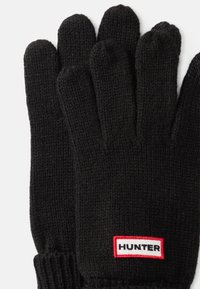 Mânuși negre tricotate cu manșetă ribbed. Prezintă o etichetă albă și roșie cu „HUNTER” brodat în zona încheieturii. Textură moale.