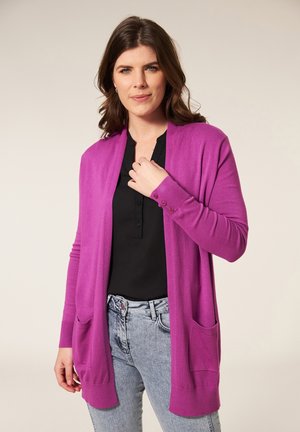 Paarse cardigan met lange mouwen, open voorkant, twee voorkruiden en knoopaccenten op de manchetten, gedragen over een zwarte top en jeans.