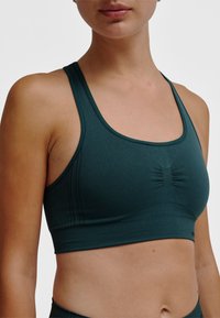 Turquoise sportbeha met naadloos ontwerp, geribbelde textuur, brede bandjes en een geplooid detail aan de voorkant. Gladde stofafwerking.