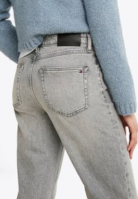 Jeans en denim gris à coupe droite, avec des détails sur les poches arrière et une étiquette en cuir noir au niveau de la taille. Portés avec un pull bleu clair.