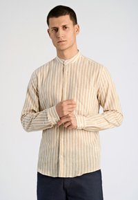 Camicia a maniche lunghe color beige chiaro con sottili righe verticali bianche, colletto a banda e tasca frontale. Sono visibili i bottoni lungo il davanti e nei polsini.