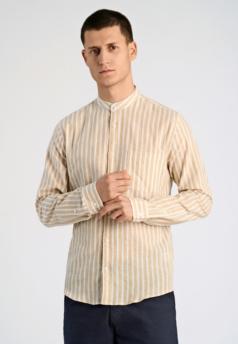 Camicia a maniche lunghe color beige chiaro con sottili righe verticali bianche, colletto a banda e tasca frontale. Sono visibili i bottoni lungo il davanti e nei polsini.