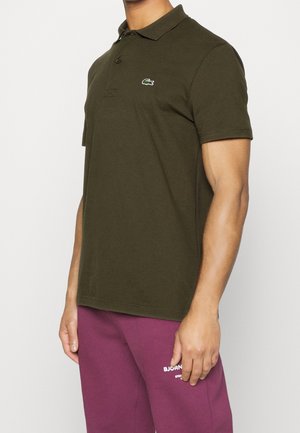 Un homme porte un polo vert foncé avec un petit logo crocodile et un pantalon de jogging violet, debout devant un fond clair et uni.
