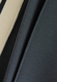 Stofprøve i beige, sort og mørkegrå, der viser glatte og teksturerede materialer med pæne kanter og varierende strikkemønstre.
