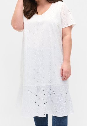 Vrouw draagt een witte, gestructureerde korte mouw jurk met geometrische uitsnijdingen over een blauwe spijkerbroek tegen een effen achtergrond.