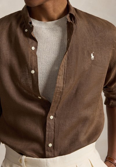 Bărbat purtând o cămașă maro din in descheiată peste un pulover alb texturat, asortată cu pantaloni albi și o brățară maro din piele la încheietură.