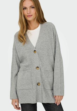Femme portant un cardigan gris clair oversize avec de gros boutons sur un haut blanc et un pantalon noir devant un fond uni.
