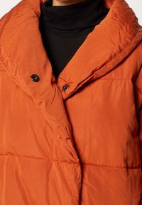 Puffig orange jacka med en stor huva, två svarta knappar och en mjuk textur, delvis överlappande framtill.