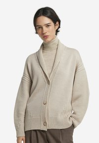 Cardigan en tricot beige avec col châle, présentant une fermeture à boutons, des poignets côtelés et de subtils détails texturés le long de l'ourlet et des épaules.