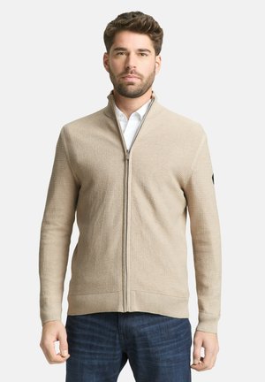 Homme portant un pull zippé beige texturé par-dessus une chemise blanche et un jean bleu foncé, debout devant un fond clair uni.