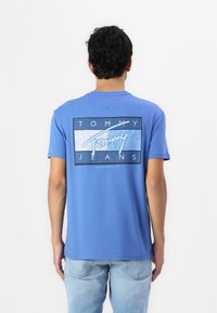 Mėlyna medvilninė marškinė su grafiniu dizainu nugaroje, kuriame yra užrašas "TOMMY JEANS", apvyniotas šviesiai ir tamsiai mėlynomis juostelėmis. Trumpomis rankovėmis.