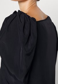 Svart blus med en design av sammanknuten puffärm, jämn textur och rund halsringning, som visar en enkel och elegant silhuett.