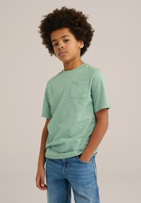 Mentolově zelené tričko s krátkým rukávem a kulatým výstřihem, s kapsou na prsou, vyrobené z měkké látky. Spárované s světle modrými džíny.