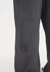 Szare spodnie z haftowanymi wzorami, w tym płomieniami i tekstem. Miękka tekstura, slim fit, z wyraźnymi wzorami po bokach.