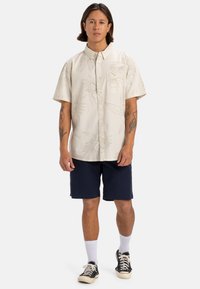 Camicetta beige a maniche corte con bottoni e motivo floreale, abbinata a pantaloni corte blu navy. Il modello indossa calze bianche a crew e sneakers nere.