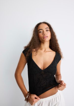 Femme aux cheveux bouclés portant un haut noir ajusté à col en V et un pantalon blanc, ajustant l'ourlet de son haut sur un fond blanc uni.