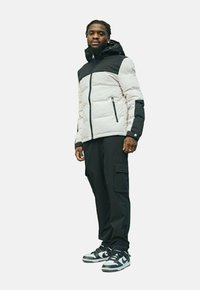 Veste d'hiver isolée avec une section supérieure noire et une section inférieure gris clair, complétée par un pantalon cargo noir et des baskets noir et blanc.