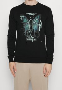 Felpa nera con collo rotondo e maniche lunghe. Presenta un design grafico con ali di farfalla e fulmini in tonalità di verde acqua e bianco.