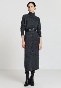 Robe tricotée gris ajustée et longue avec un col roulé haut et des manches longues, cintrée à la taille par une ceinture en cuir noir. Bottes noires à talons.