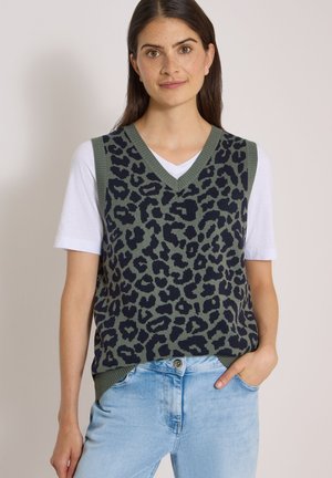Femme portant un pull sans manches imprimé léopard vert et noir sur une chemise blanche à manches courtes avec un jean bleu clair, main dans la poche.