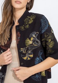 Musthave lühikeste varrukatega jakki, millel on mitmevärviline lilleline tikand, väljaulatuva kraega ja mustad nööbid, tekstureeritud kootud kangas.