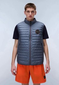 Jovem vestido com um colete cinza acolchoado sobre uma camisete preta de manga curta e calções laranja brilhante, posando contra um fundo simples.