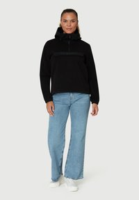 Sort fleece pullover med halv lynlås, justerbar hætte og bånd over brystet. Kombinationen er med lyseblå bukser med bred benvidde og hvide sneakers.
