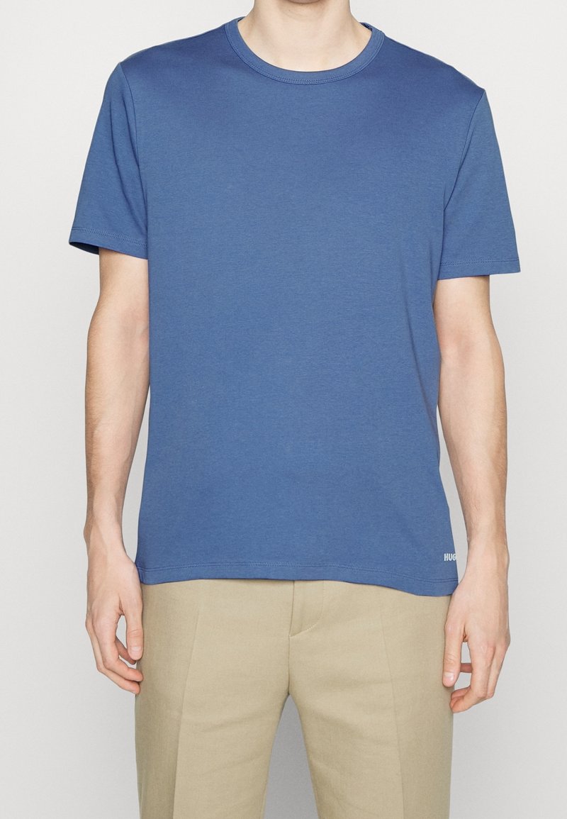 Homme portant un t-shirt bleu uni à manches courtes et col rond, et un pantalon beige, debout avec les bras détendus le long du corps.