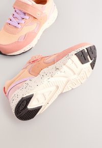 Buty sportowe w różowo-lawendowej kolorystyce, posiadające teksturowaną cholewkę, pasek na rzep oraz gumową podeszwę w cętki z wyraźnym wzorem bieżnika.