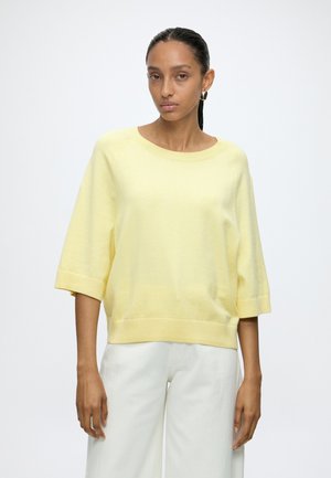 Donna con capelli scuri raccolti, che indossa orecchini a cerchio, un maglione largo giallo chiaro e pantaloni bianchi su uno sfondo chiaro uniforme.