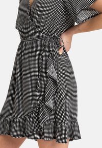 Musta-valkoinen ruudullinen lyhyt kietaisumekko röyhelöhemmoilla, nauhoitettu vyötärö ja lyhyet hihat, päällä henkilö, jonka käsi on lanteella.