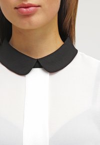 Biała koszula z czarnym, zaokrąglonym kołnierzykiem; gładki materiał; centralny pionowy szew. Skupienie na designie kołnierza i kontraście materiałów.