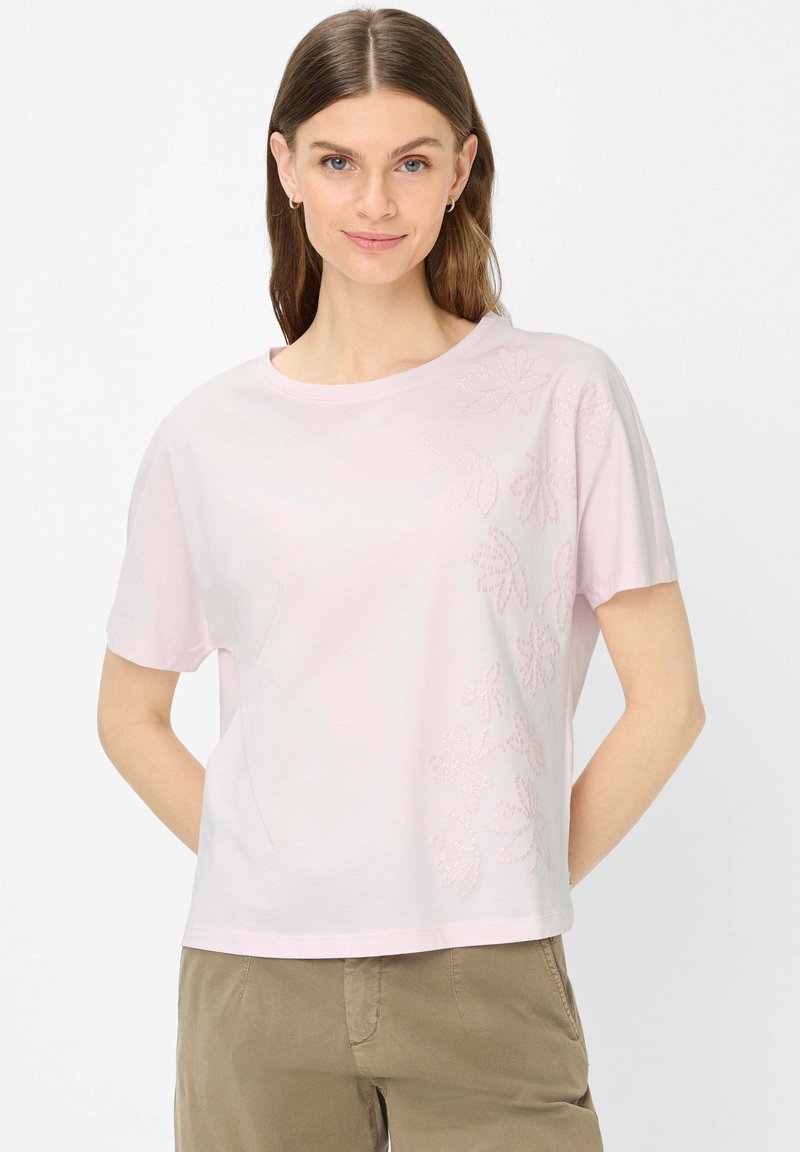 Femme portant une chemise à manches courtes rose clair avec une broderie florale subtile sur le côté droit, associée à un pantalon beige, debout les mains derrière le dos.
