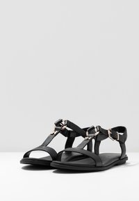 Svarta lädersandaler har en öppen tådesign med korsade remmar. Anmärkningsvärda spännen i guldton pryder anklarremmarna på en platt sula.