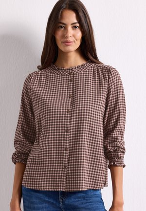 Femme aux longs cheveux foncés portant un chemisier à boutons à carreaux marron et rose avec des poignets froncés et un jean bleu, debout contre un mur clair.
