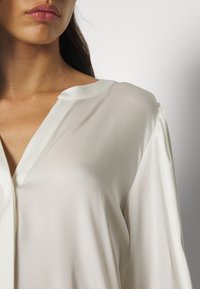 Seidenweiße Bluse mit tiefem V-Ausschnitt, dezentem Schimmer und lockeren Ärmeln. Sie hat einen abgerundeten Saum und minimalistische Designdetails.