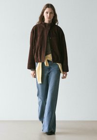 Braune Bomberjacke aus Wildleder mit hohem Kragen und Knopfverschluss, getragen über einem gestreiften Top und kombiniert mit weit geschnittenen Jeans.